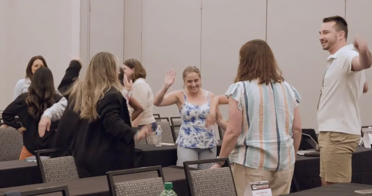 Participants smiling and reaching toward each other during a paired improv exercise in a conference room.
