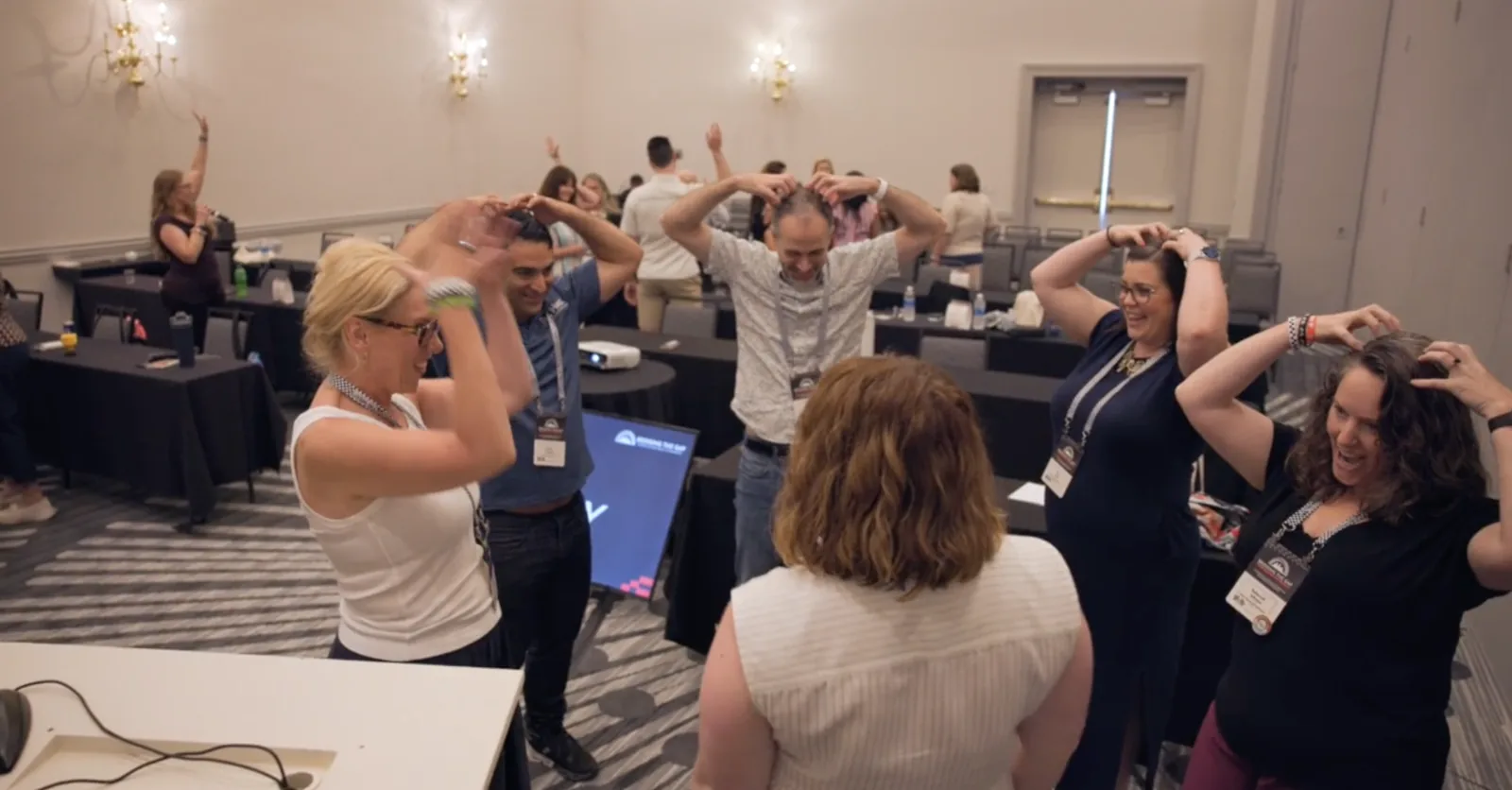 A room of workshop participants laughing together during a group warm-up game, hands held above their heads like antlers.