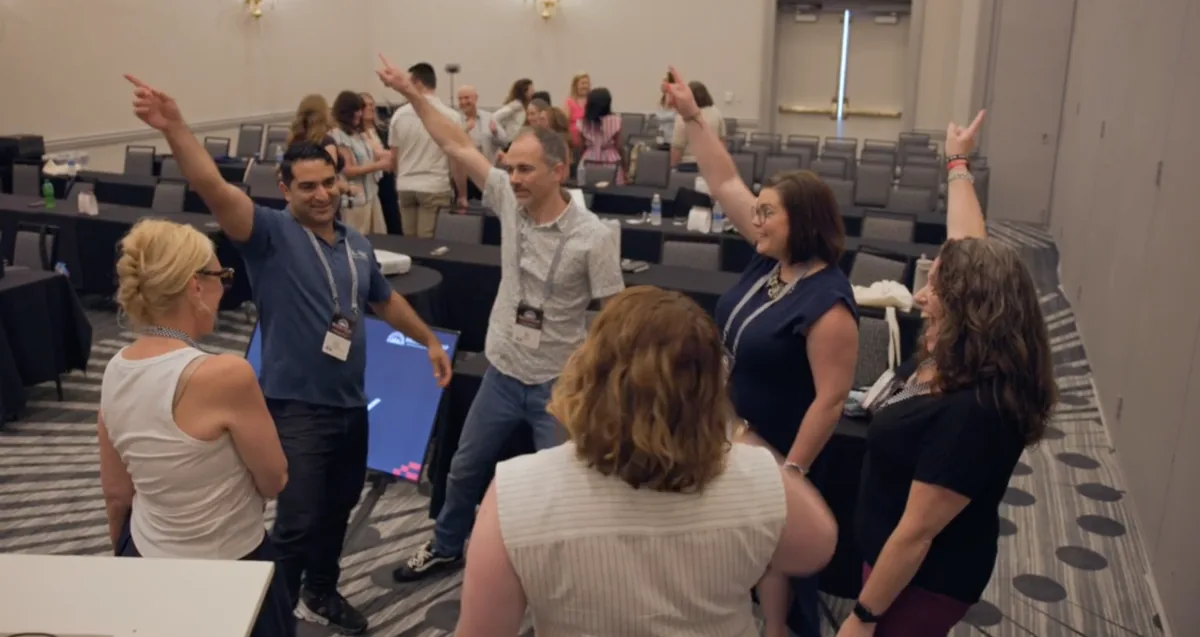 A group of workshop participants standing with arms raised and pointing, laughing during a warm-up exercise.