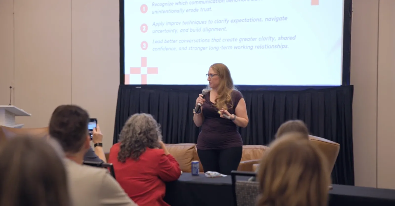 Jennifer Dymond speaking with a microphone on a conference stage, in front of a slide listing the session&rsquo;s learning objectives.